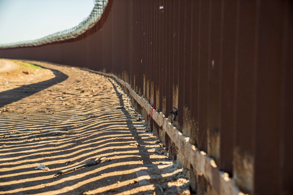 Photo of the border wall curving to the left, topped with coils of barbed wire, casting shadows. 